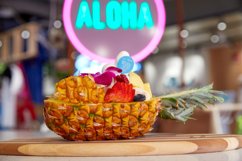 Half cut pineapple with cut strawberries, pineapple and soft serve on top and a neon green Aloha sign with a neon pink circle in the background